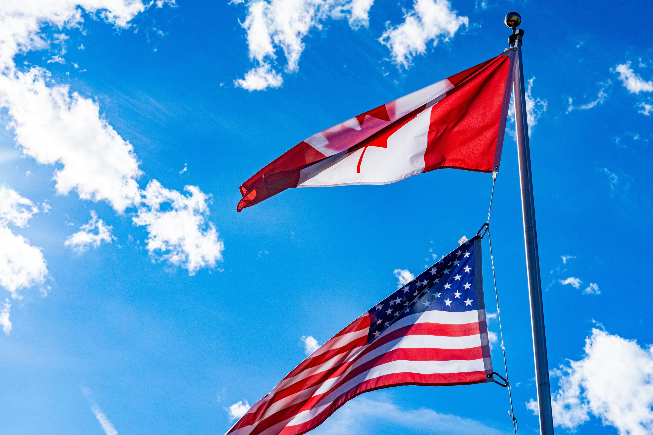 chris-robert-w0oiai6dcJM-unsplash - Edwards Law An image showing a Canadian and U.S. flag on a flagpole, blowing in the wind together with the sunny blue sky in the background.